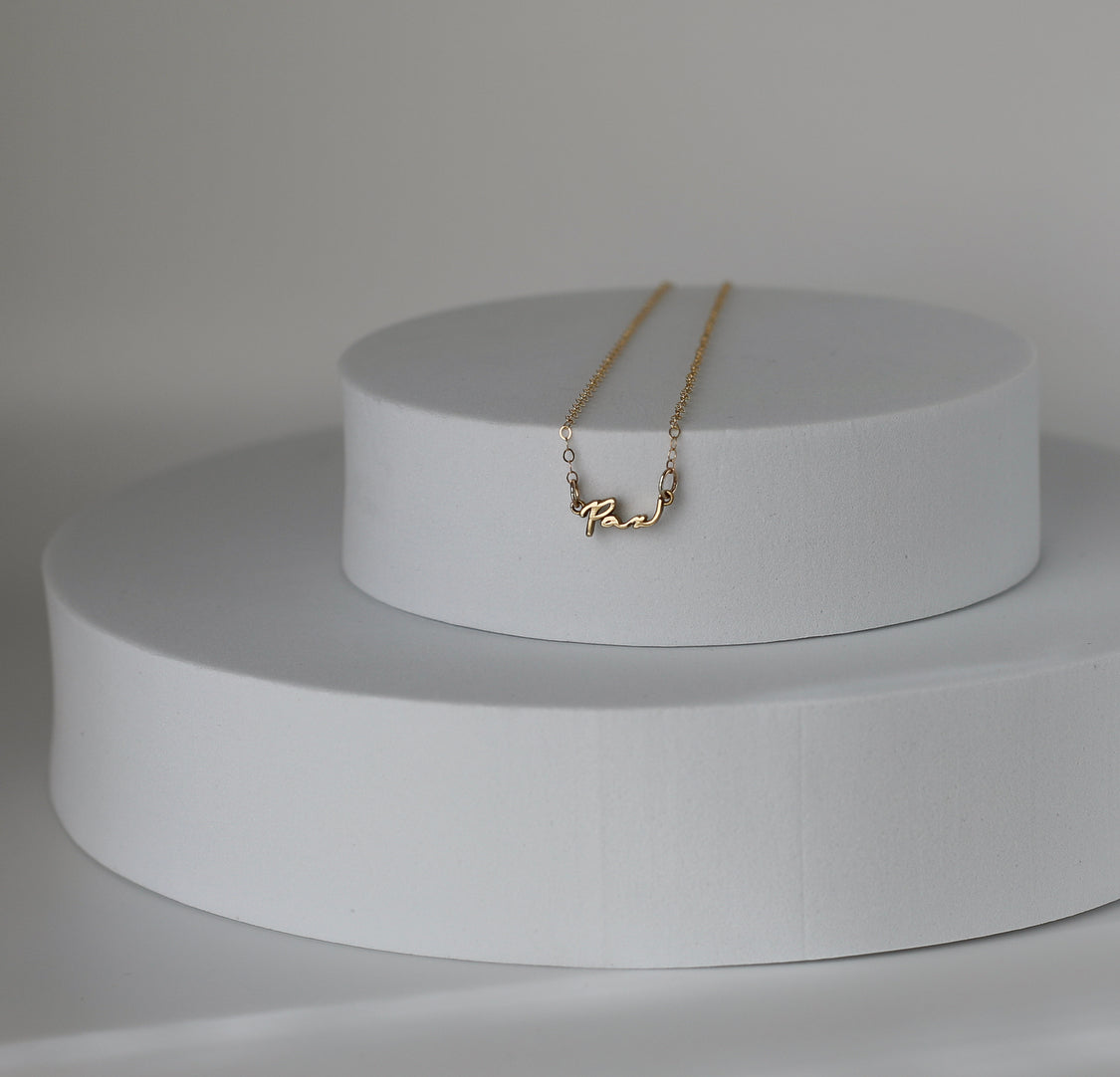 Gold Pax pendant necklace on white two-tier display pedestal