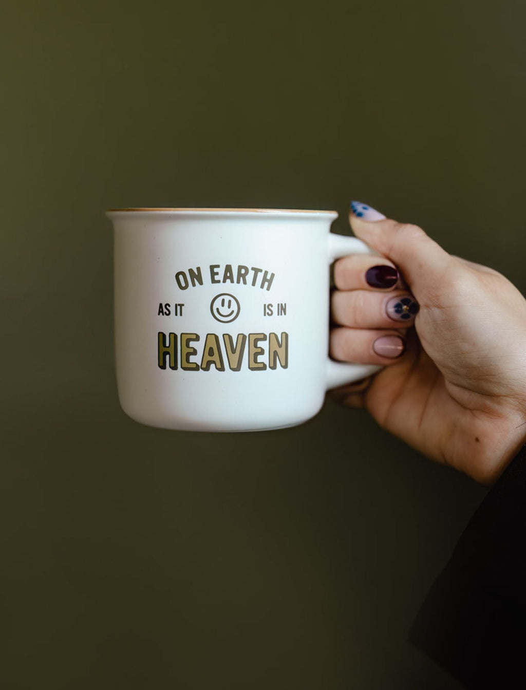 White ceramic mug held in hand, green "ON EARTH AS IT IS IN HEAVEN" text.