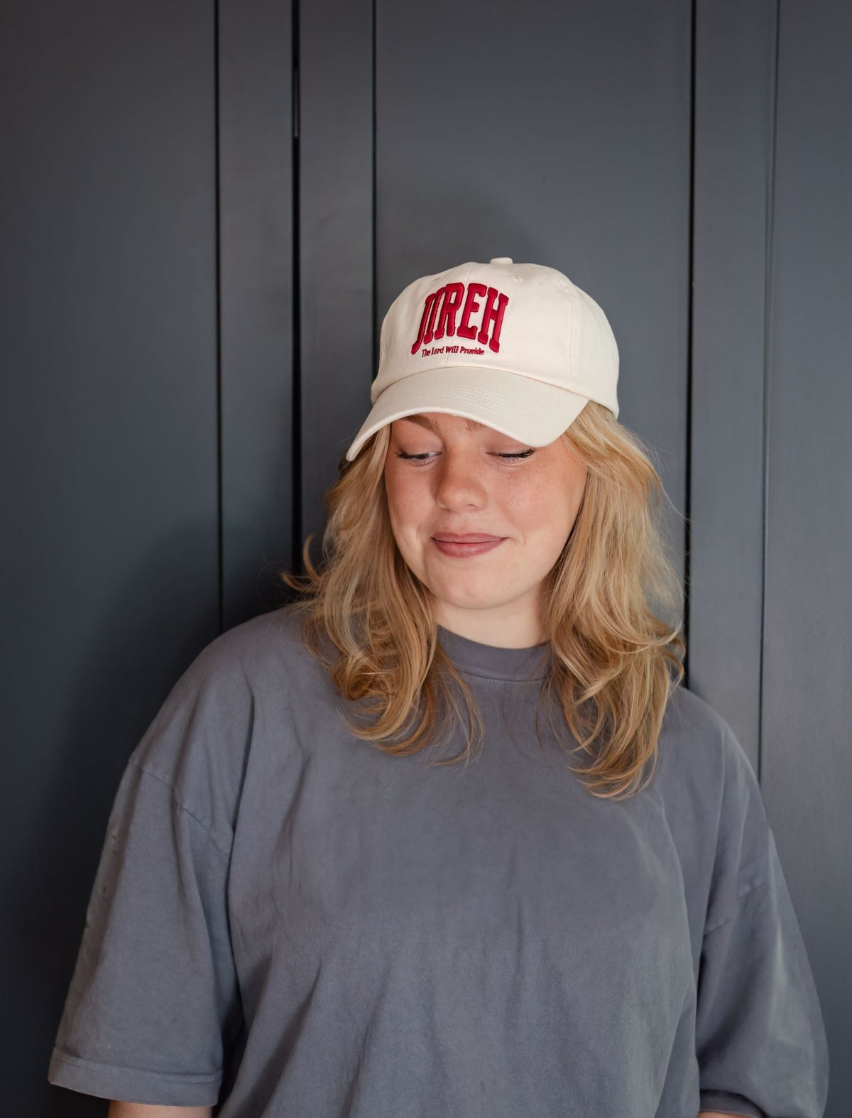 Beige baseball cap with red logo, worn with brim angled to the side