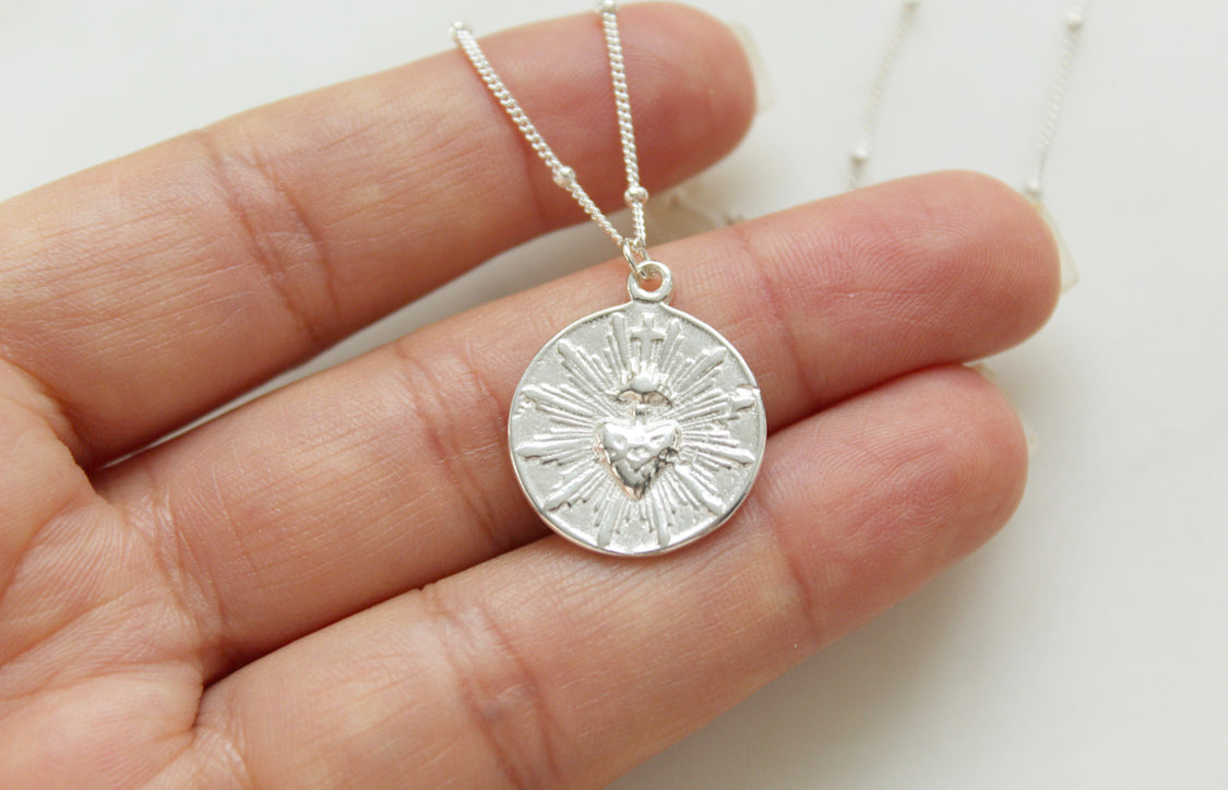Silver round sacred heart coin pendant with radiating rays on delicate chain.