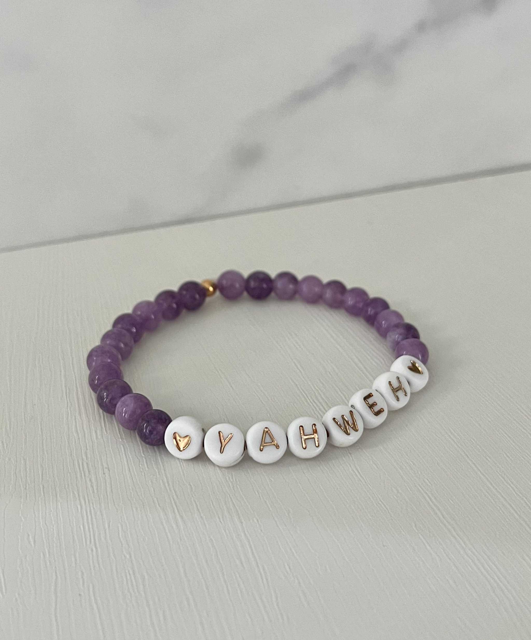 Purple beaded stretch bracelet with white letter charms spelling YAHWEH and heart details.