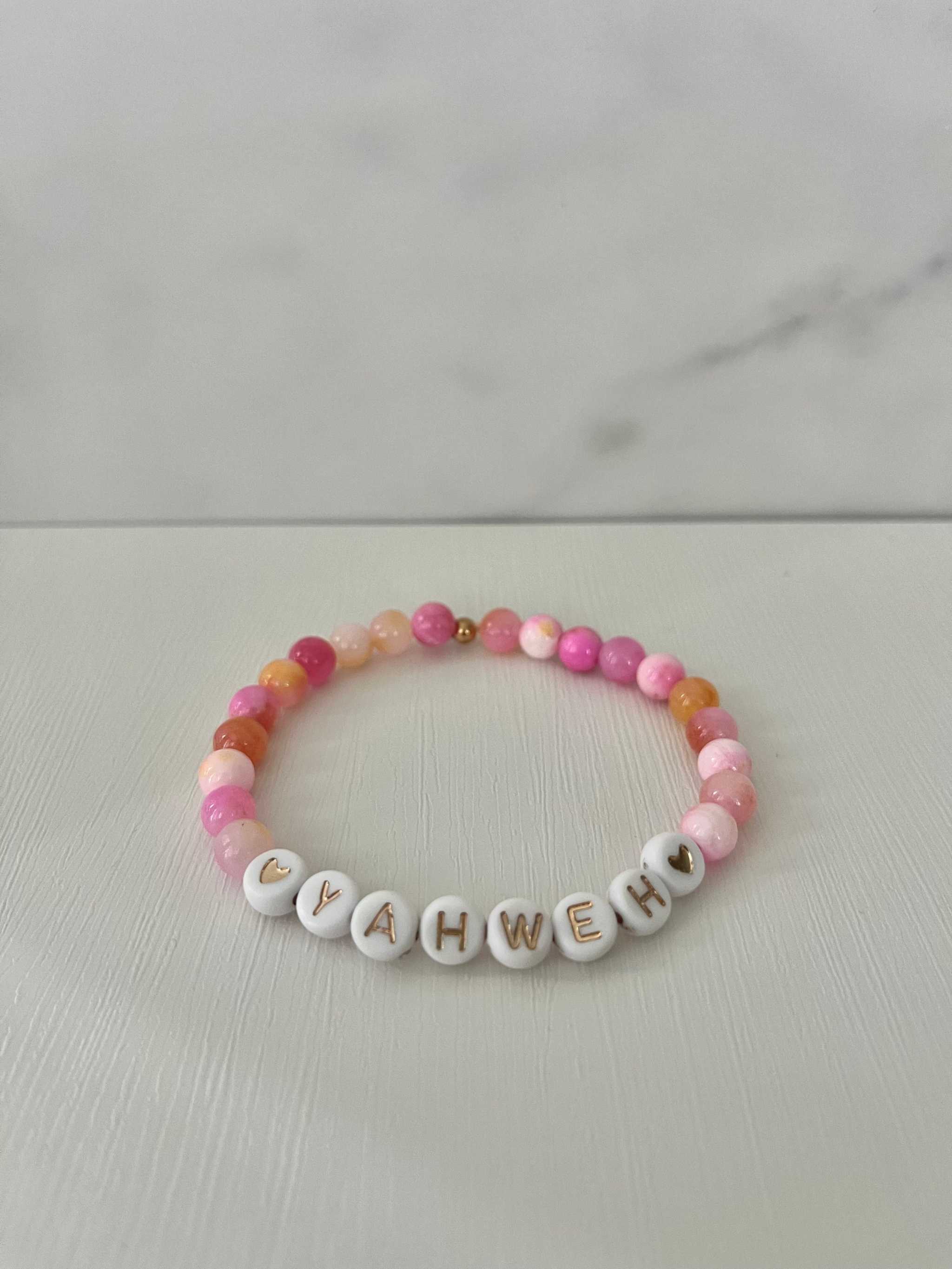 Pink beaded stretch bracelet with white letter charms spelling YAHWEH and small heart details.