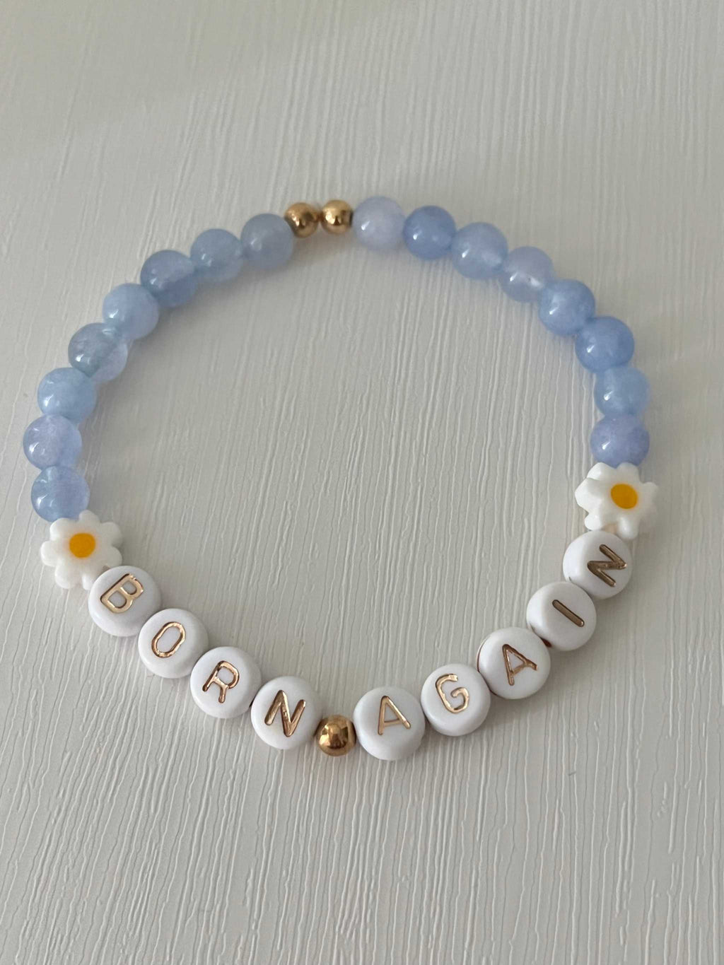 Sky blue beaded bracelet with white alphabet beads and gold spacer accents; flower charms on sides.