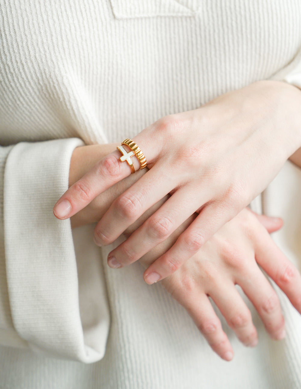 Gold spacer ring with cross charm, worn on finger against cream sweater