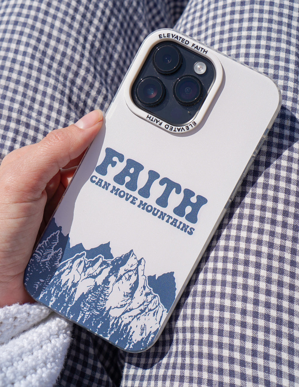 Hand holding white phone case with “FAITH CAN MOVE MOUNTAINS” text and blue mountains