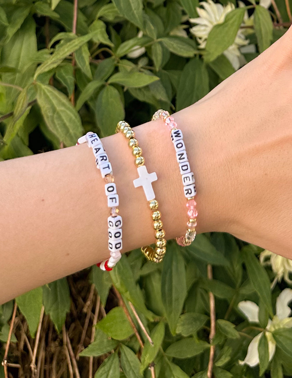Gold beaded cross bracelet with white cross charm and white letter beads on wrist