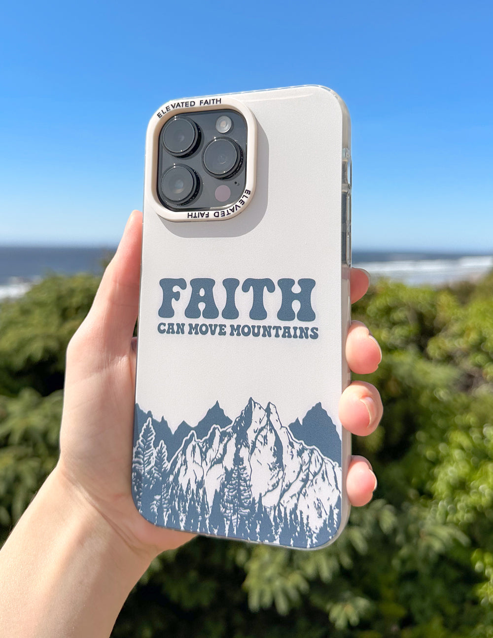 Hand holding white phone case with “FAITH CAN MOVE MOUNTAINS” and blue mountain graphic