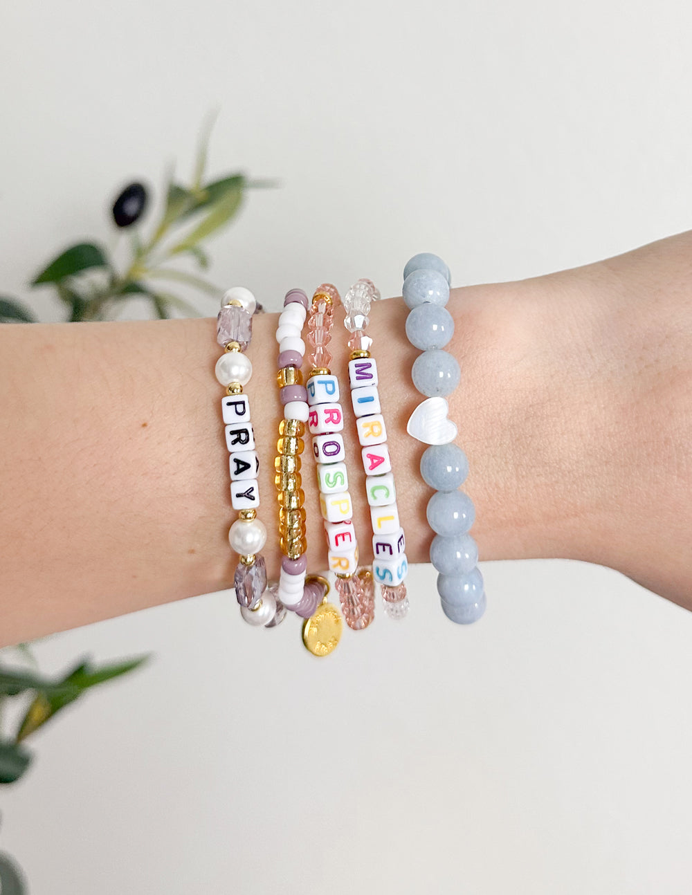 Stacked letter bracelets with beads spelling “PRAY” and “MIRACLES”, plus pearl beads.