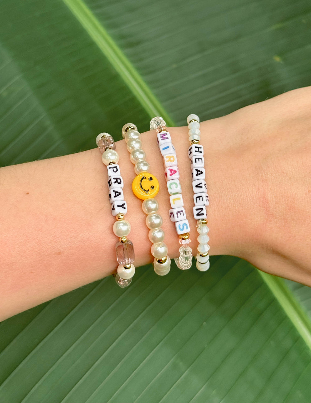 Stacked beaded letter bracelets with gold coin charm and beads spelling “HEAL”.
