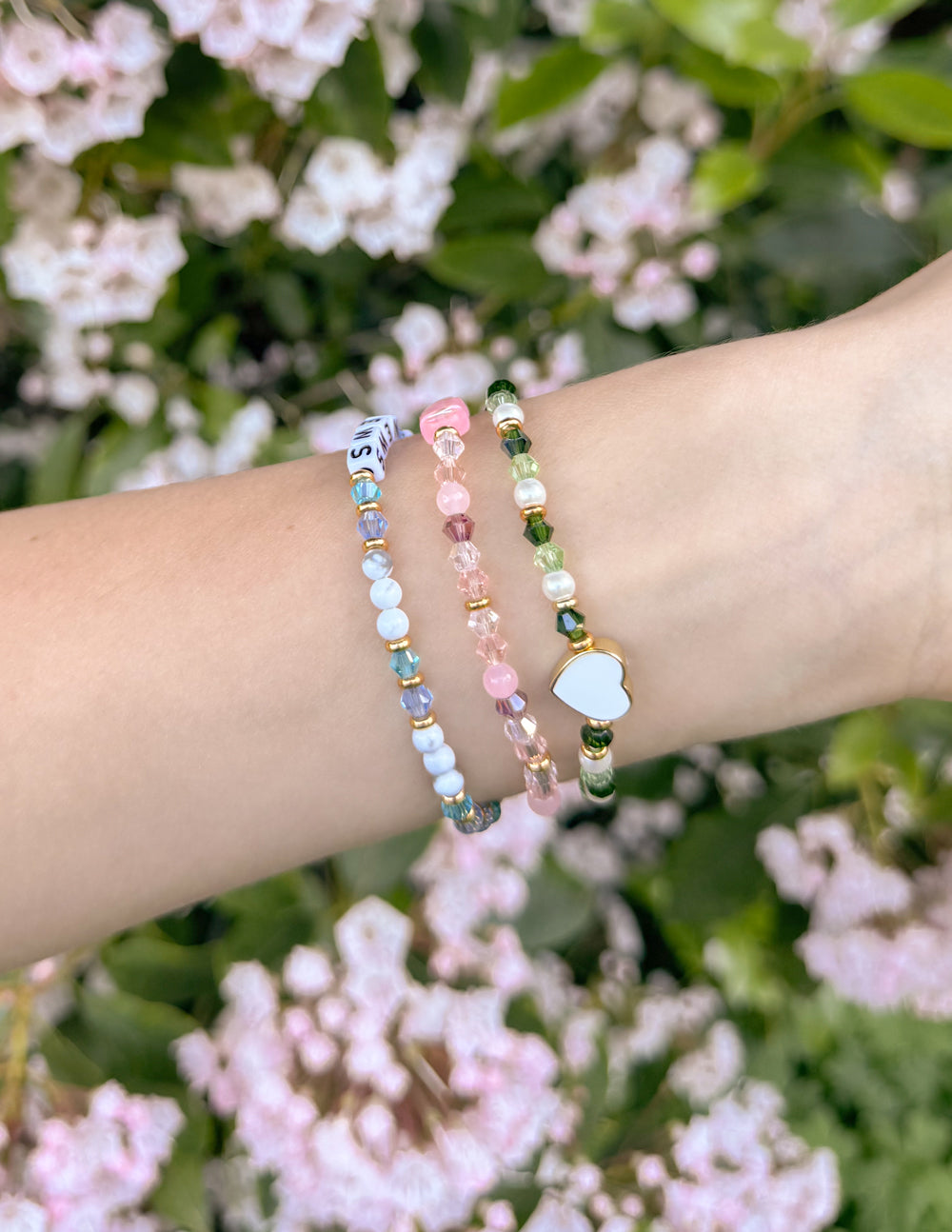 Stacked beaded letter bracelets on wrist with pink, blue, and a small green charm