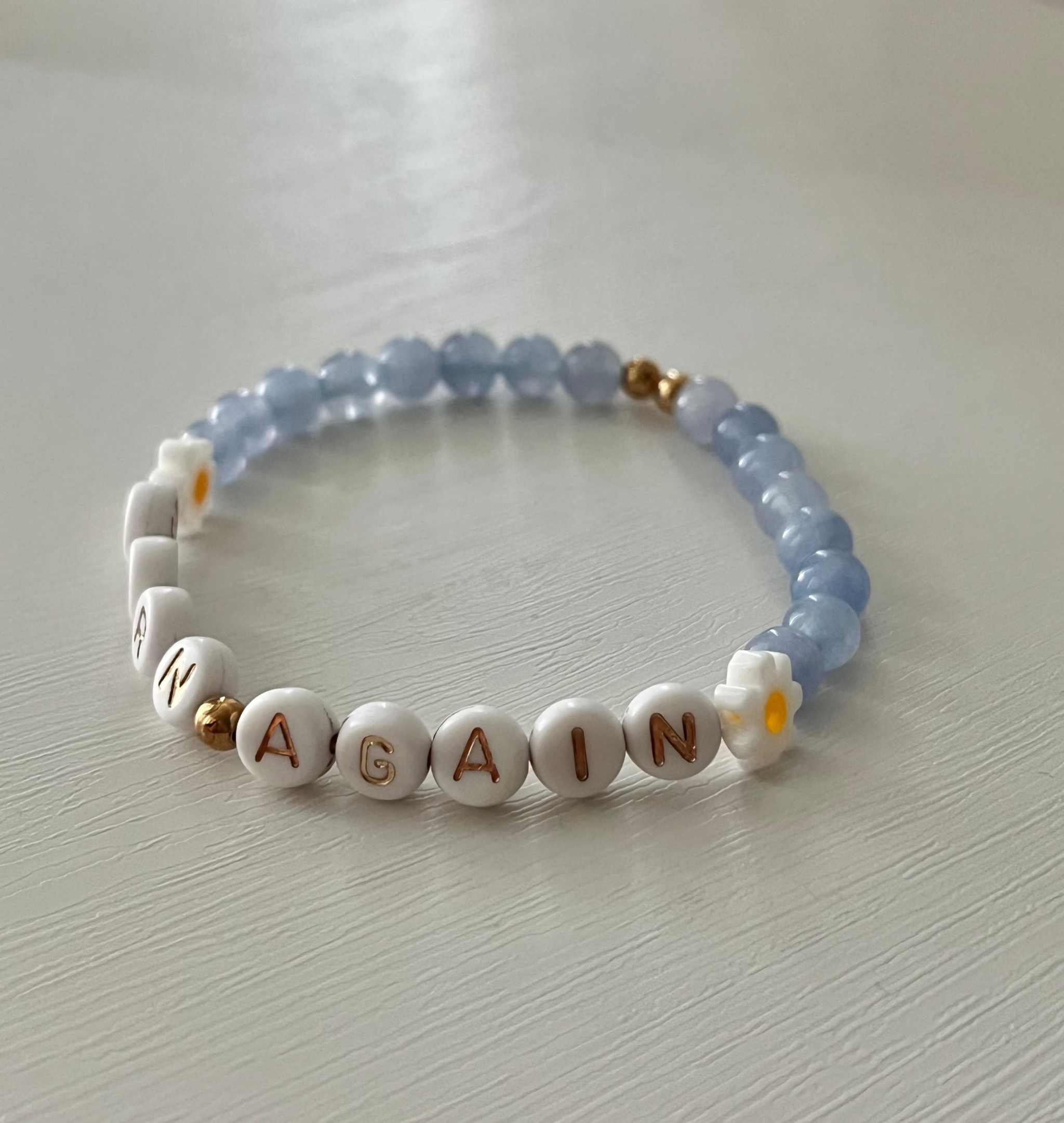 Sky blue and white beaded bracelet spelling “BORN AGAIN” with small yellow flower charms and gold bead.