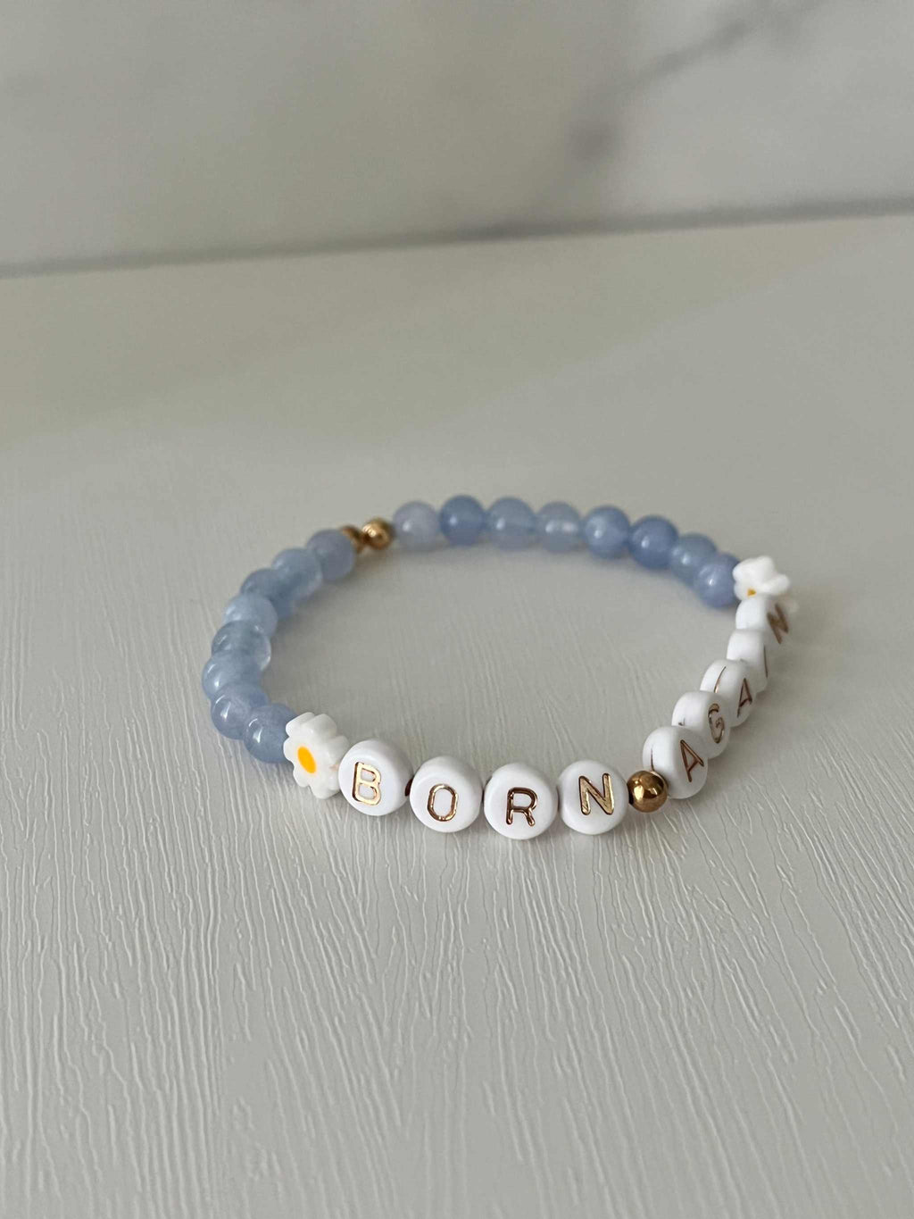 Sky blue bracelet with white letter beads spelling “BORN AGAIN” and tiny flower charms.