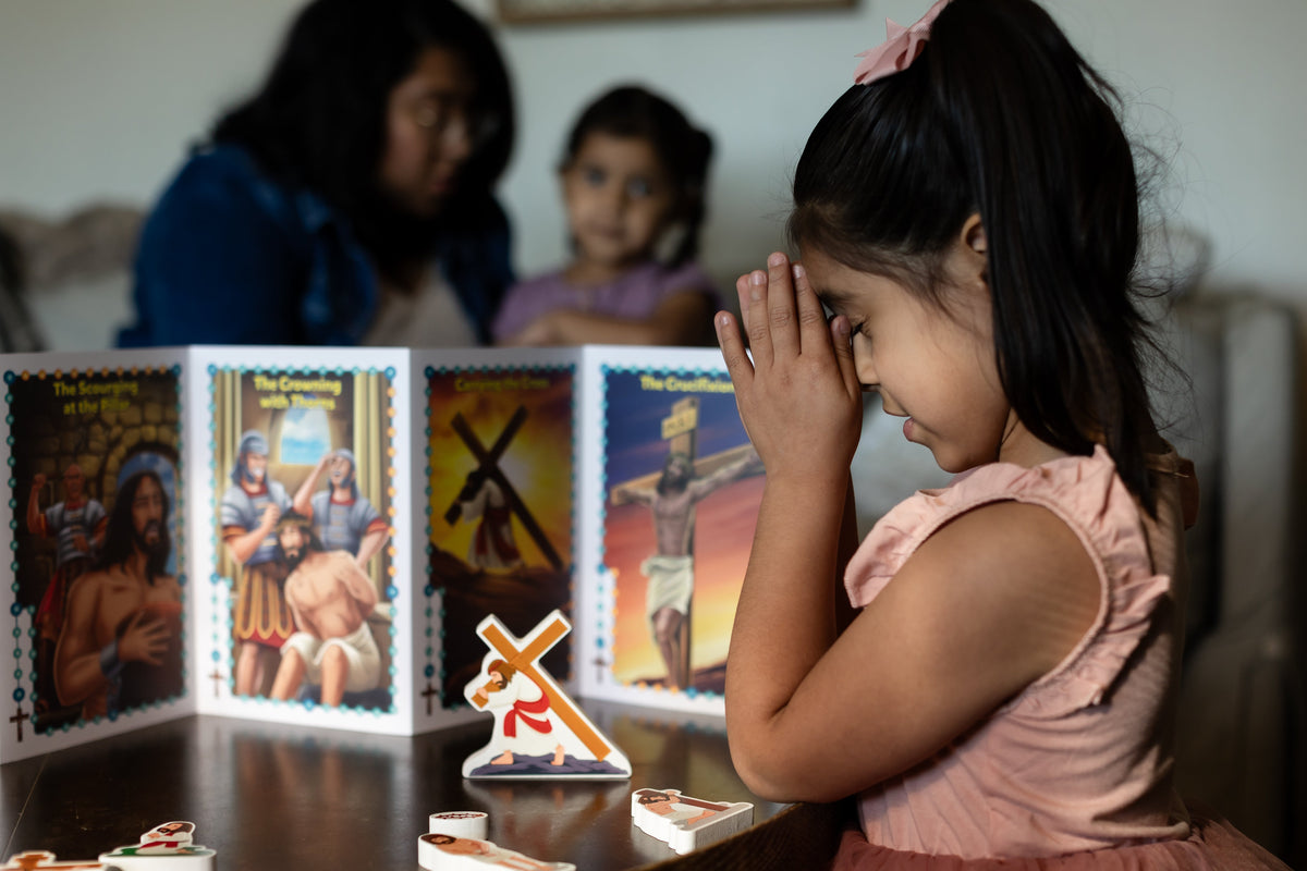 Child praying with small religious pieces beside illustrated card panels from a rosary play set