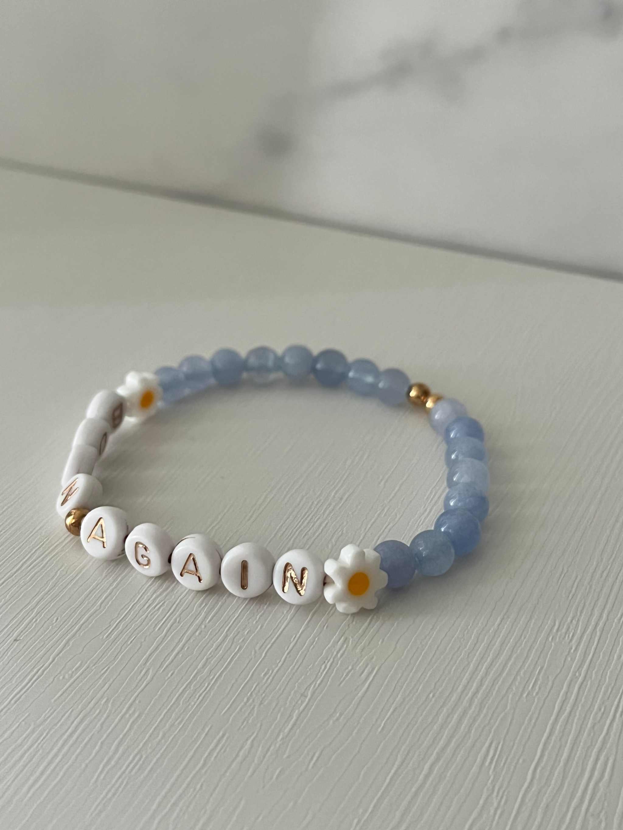 Sky blue beaded bracelet with white alphabet beads spelling “BORN AGAIN” and daisy flower details.