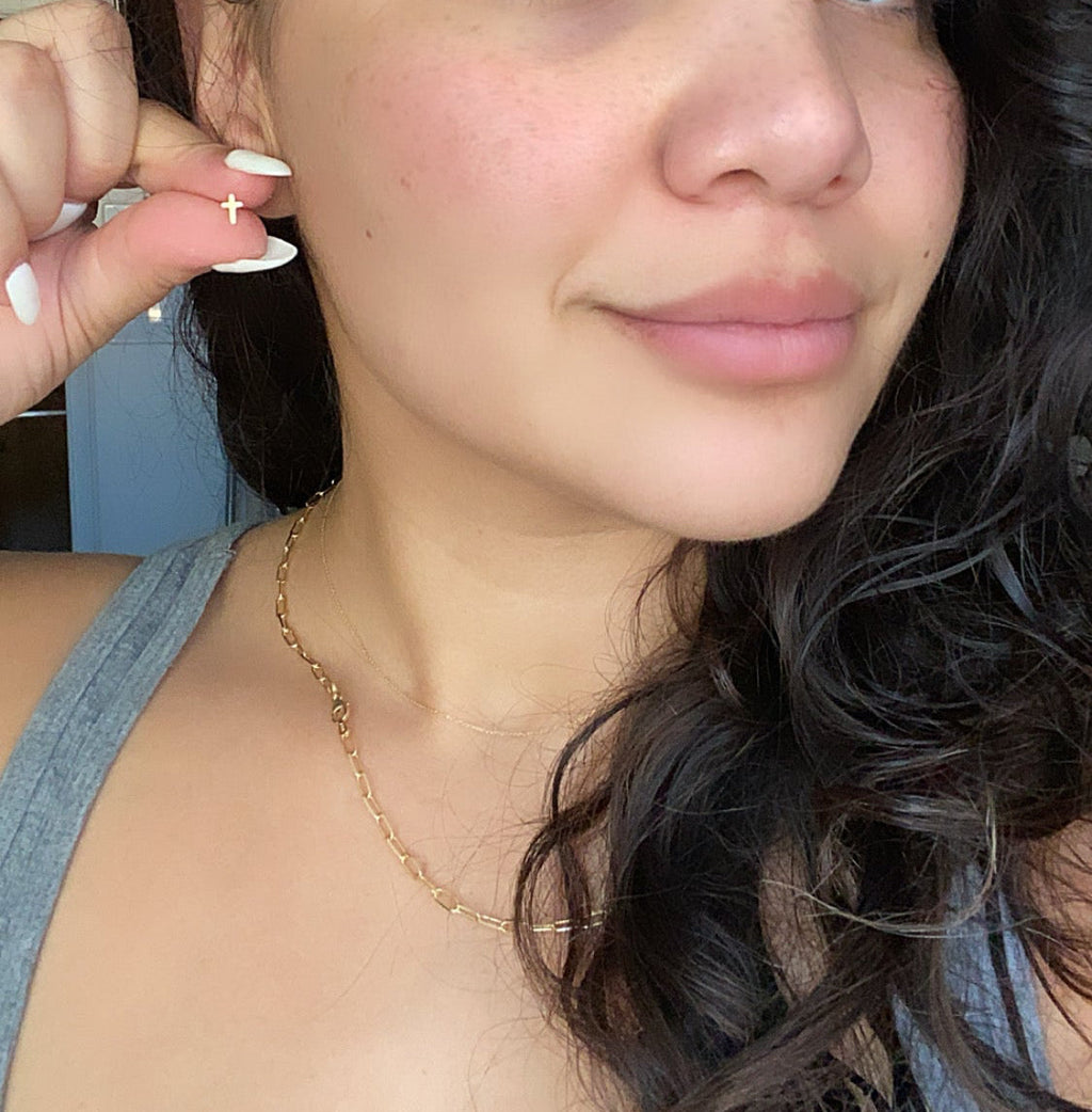 Gold-tone cross stud earrings on the ear, worn with a delicate chain necklace.