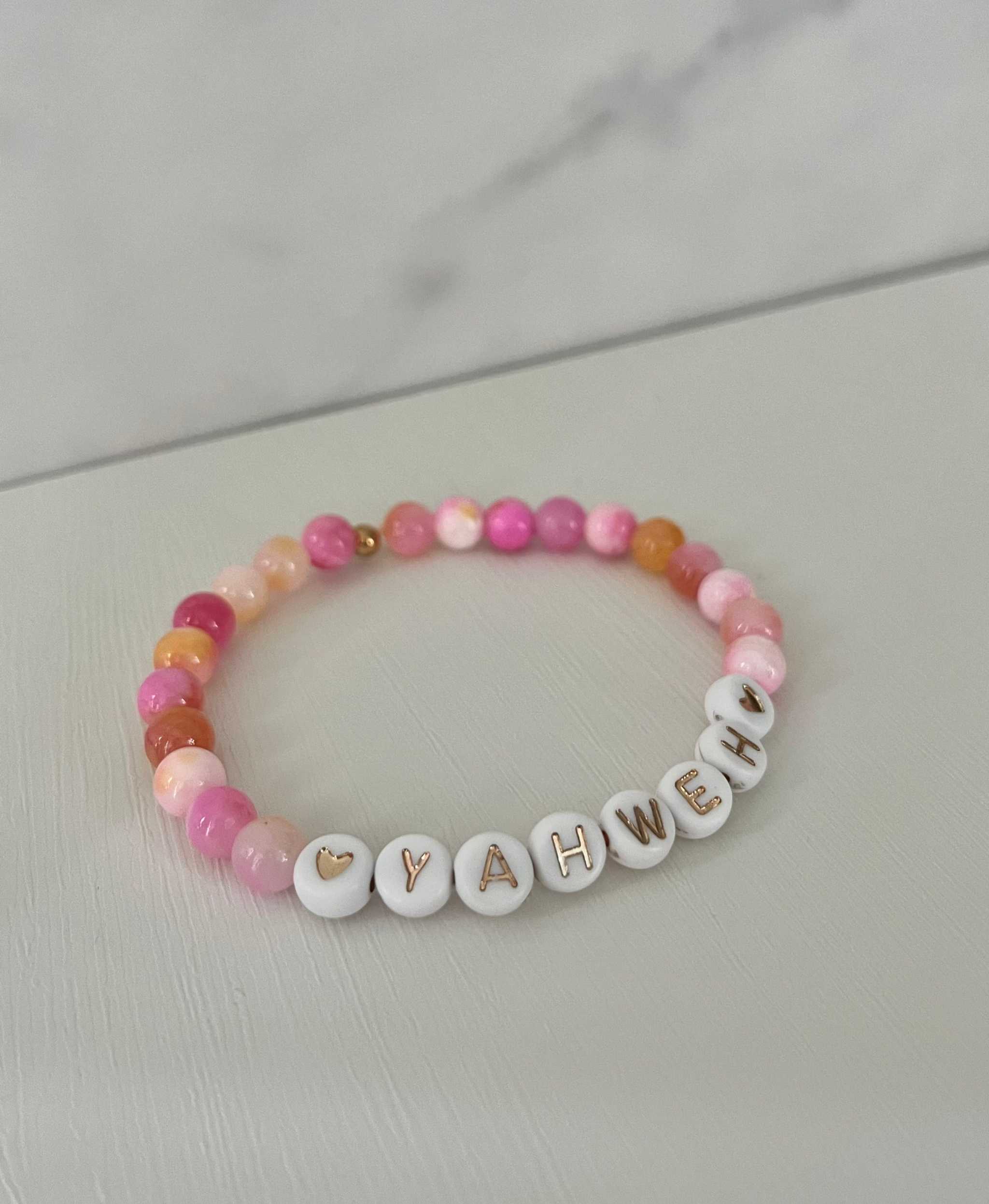 Pink beaded stretch bracelet with white letter charms spelling YAHWEH and heart details.