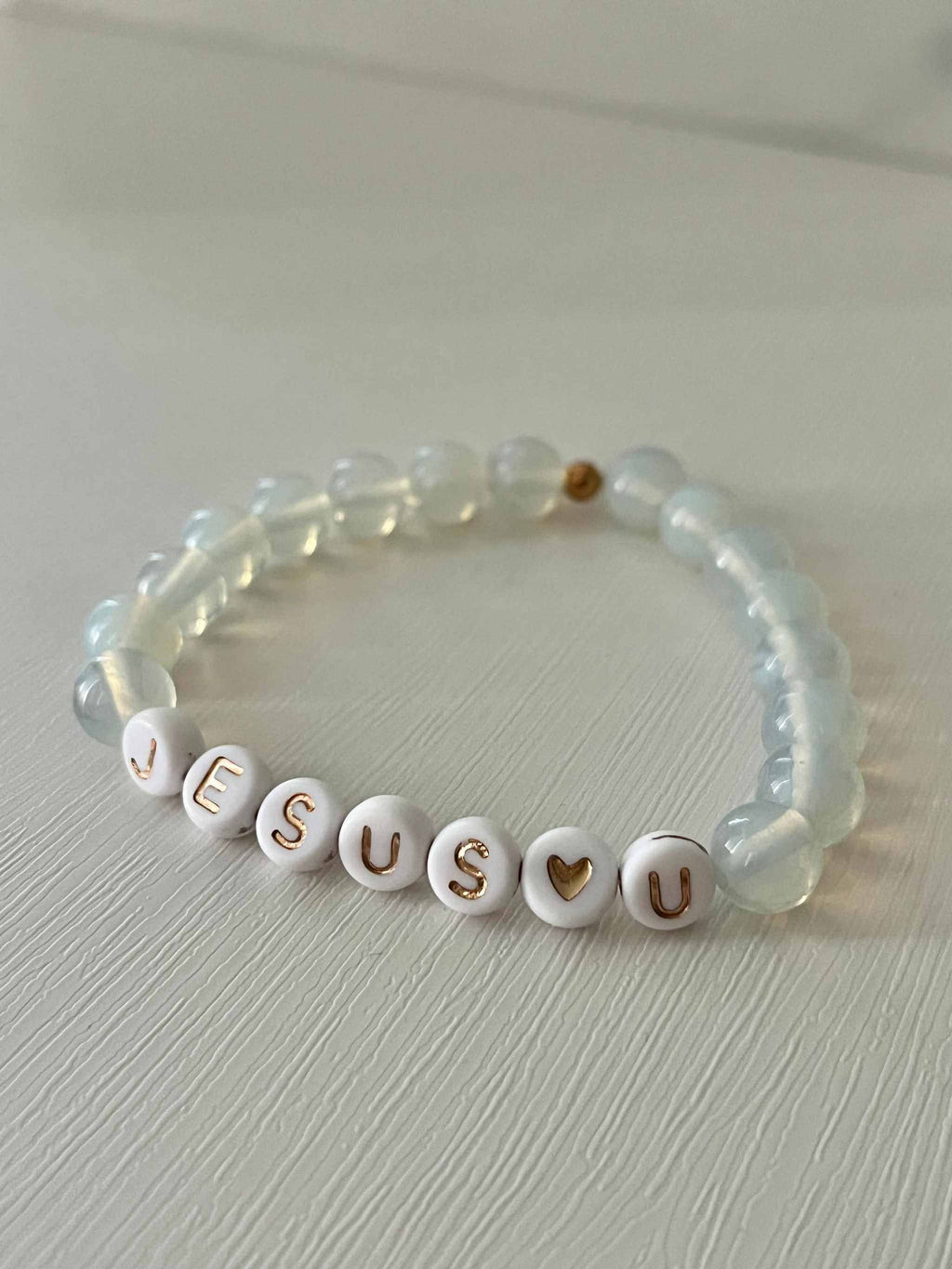 Clear bead bracelet with white letter beads spelling "JESUS U" and gold accent bead