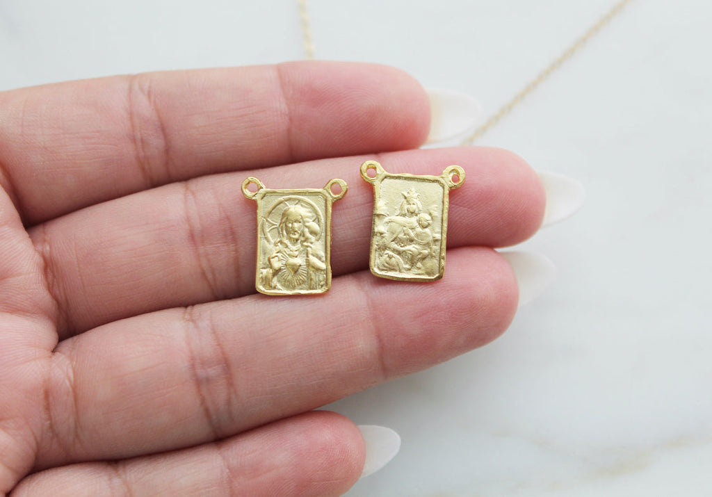 Scapular charm pair with square gold-tone frames and relief faces, shown in hand.