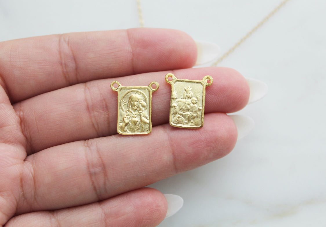 Scapular charm pair with square gold-tone frames and relief faces, shown in hand.