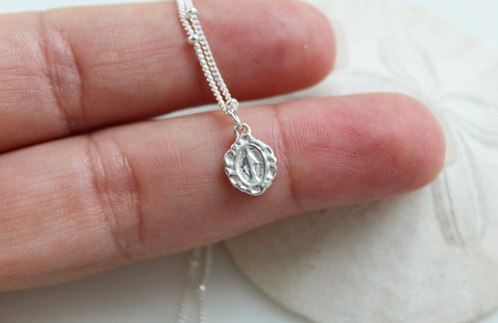 Tiny sterling silver oval medal pendant on textured chain, held above cloth