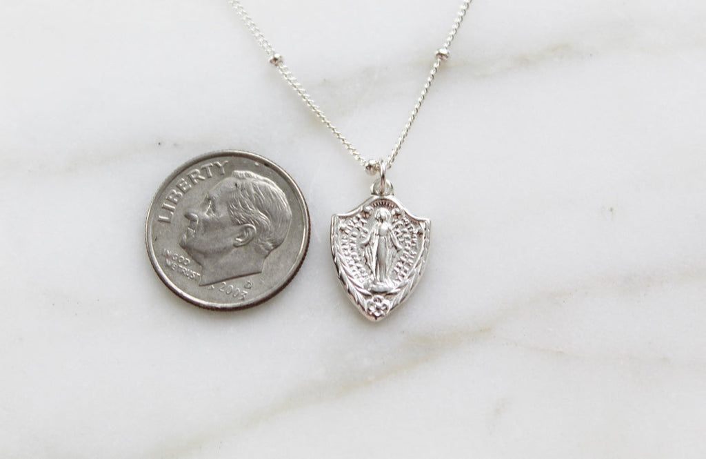 Sterling silver shield pendant necklace with Virgin Mary figure, shown with coin for scale