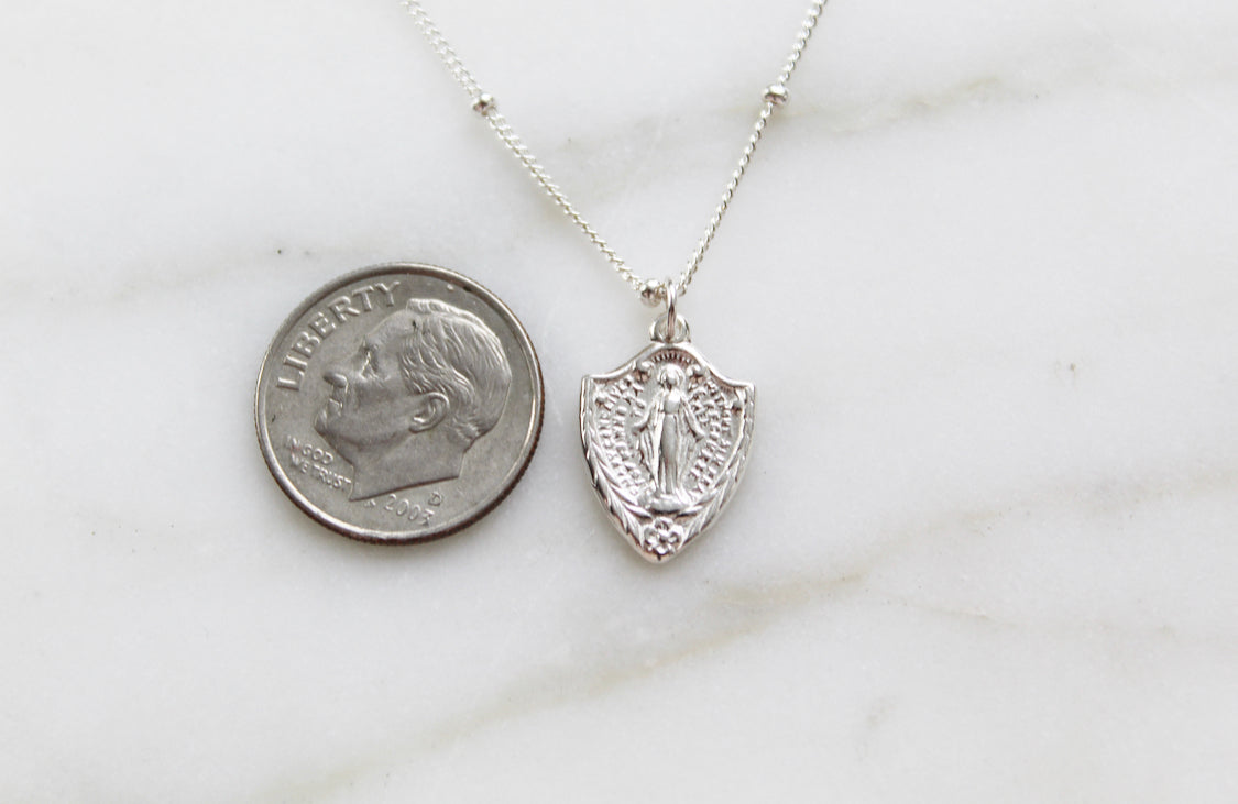 Sterling silver shield pendant necklace with Virgin Mary figure, shown with coin for scale