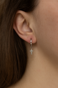 Silver huggie hoop earrings with dangling cross charm, shown on ear close-up