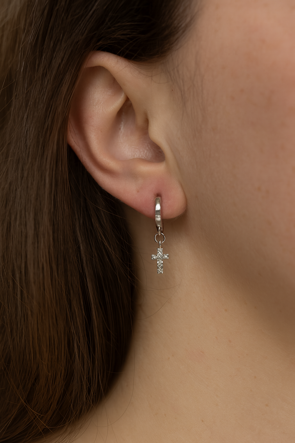 Silver huggie hoop earrings with dangling cross charm, shown on ear close-up