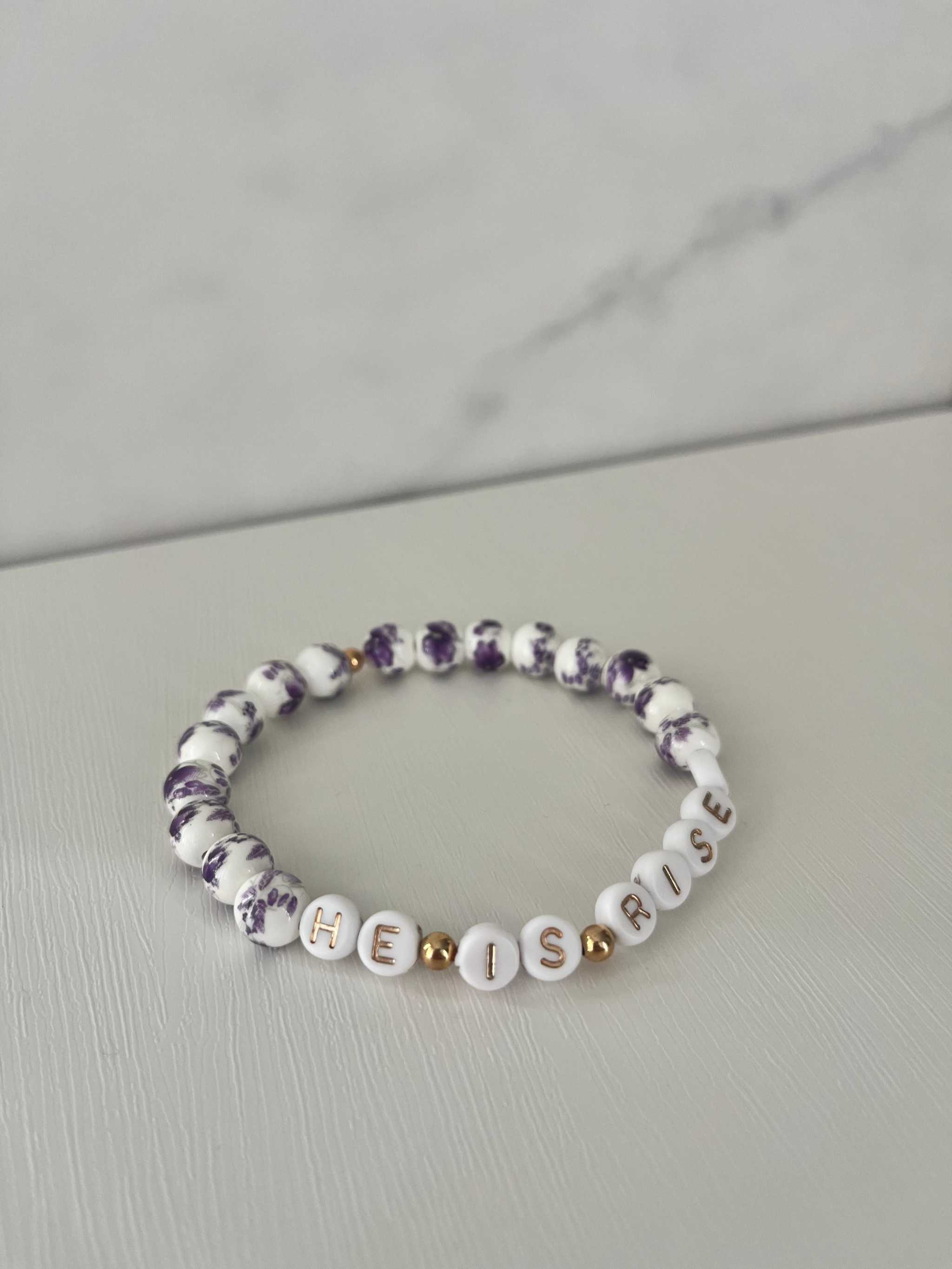 Purple and white bead bracelet spelling “HE IS RISEN” with gold cross charm