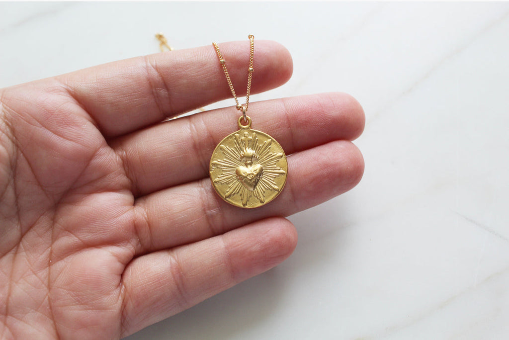 Gold-tone sacred heart coin pendant on thin chain, held in a hand
