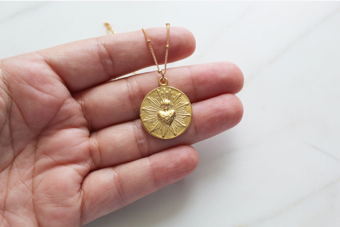 Gold-tone sacred heart coin pendant on thin chain, held in a hand