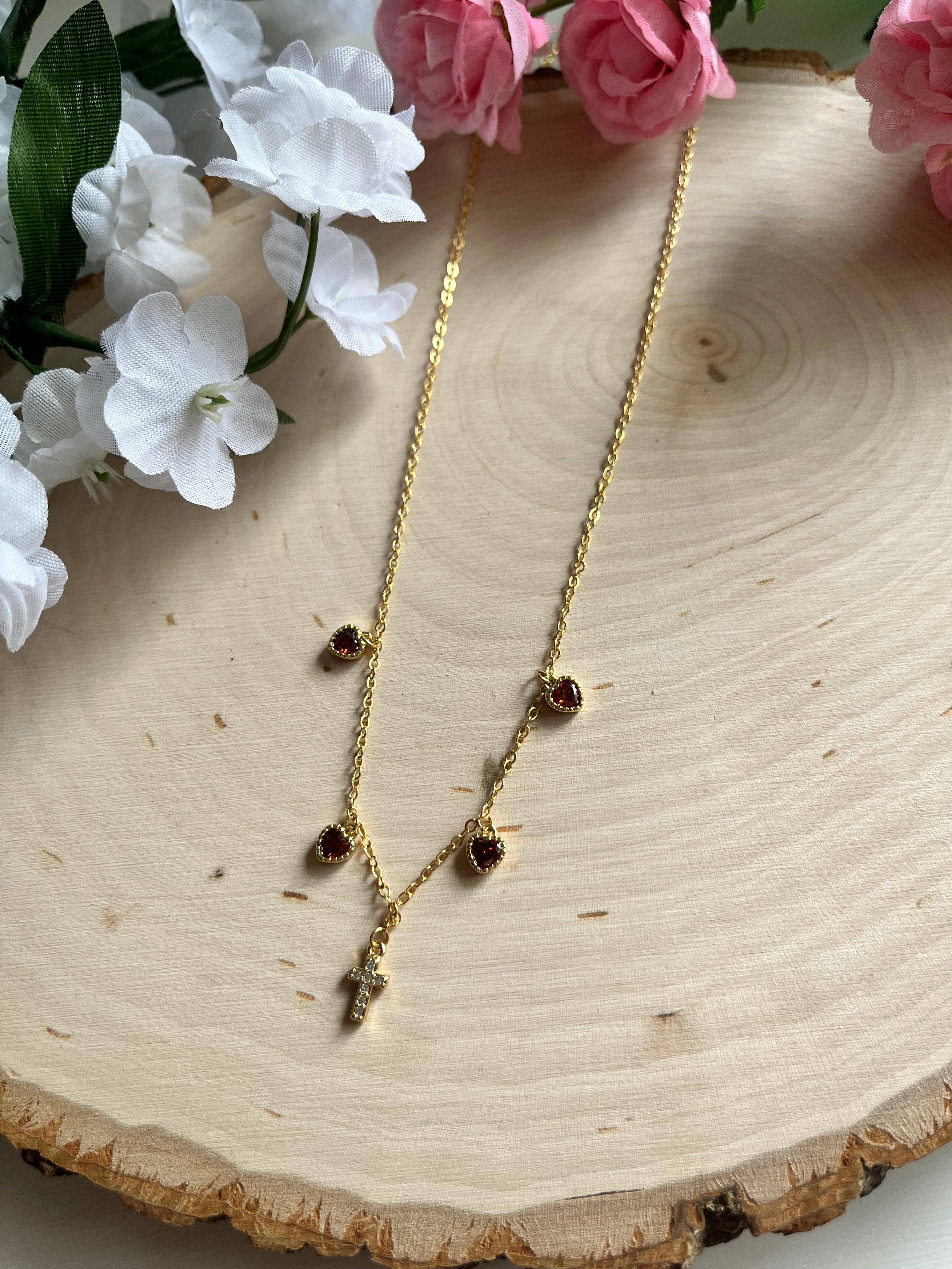 Gold charm necklace with red heart stones and gold cross pendant, close angled view