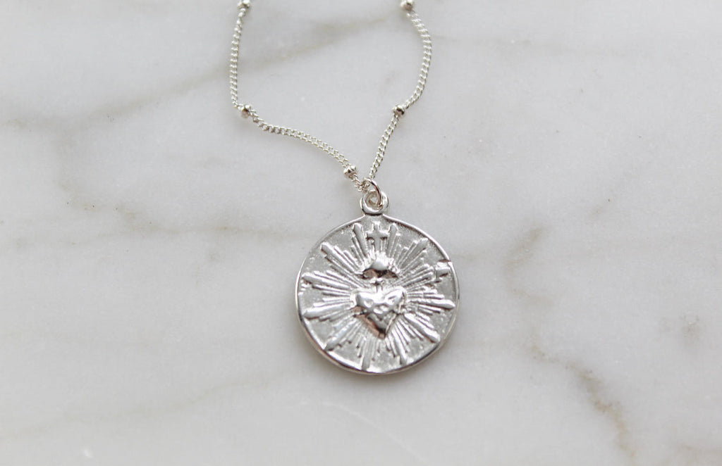 Silver round sacred heart coin pendant necklace on a white marble surface.
