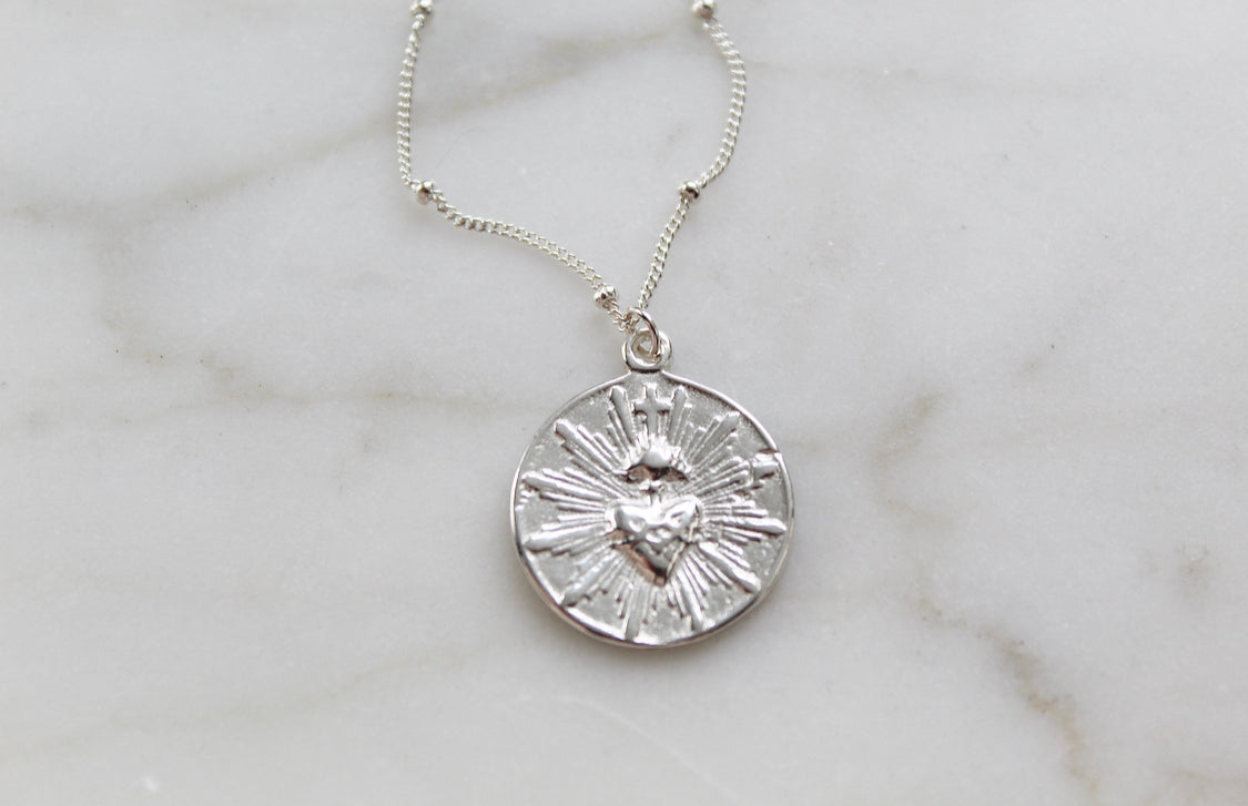 Silver round sacred heart coin pendant necklace on a white marble surface.