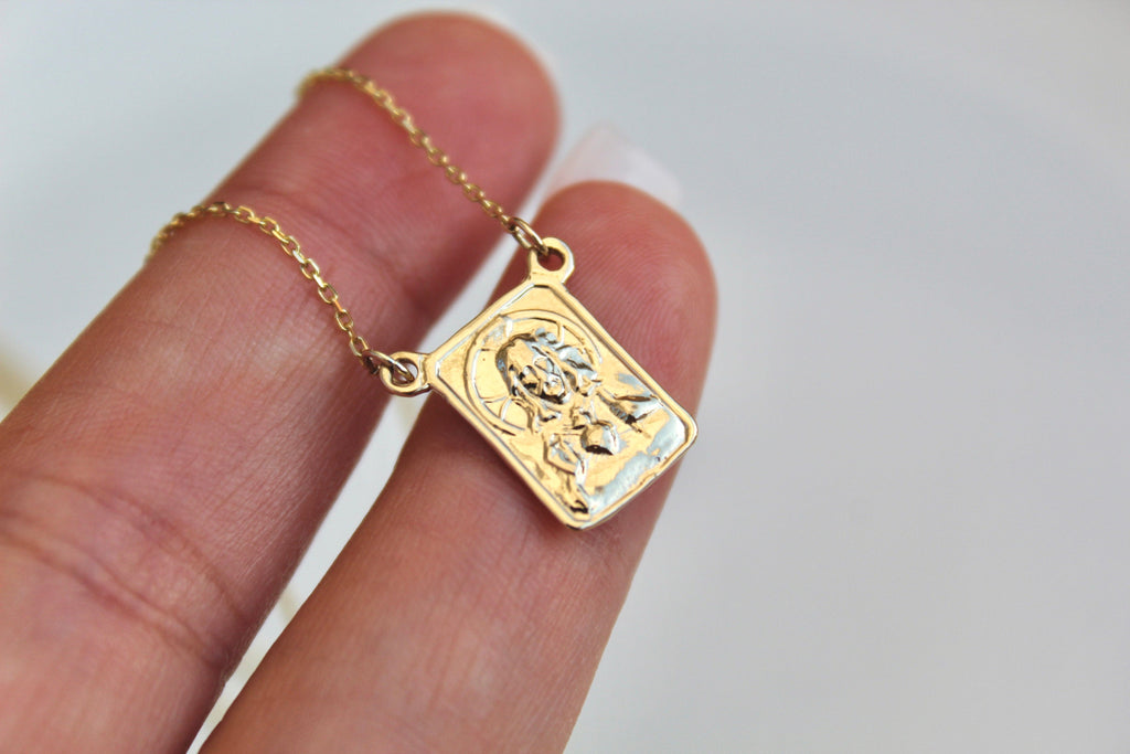 Gold scapular pendant on chain held by fingers, square embossed front