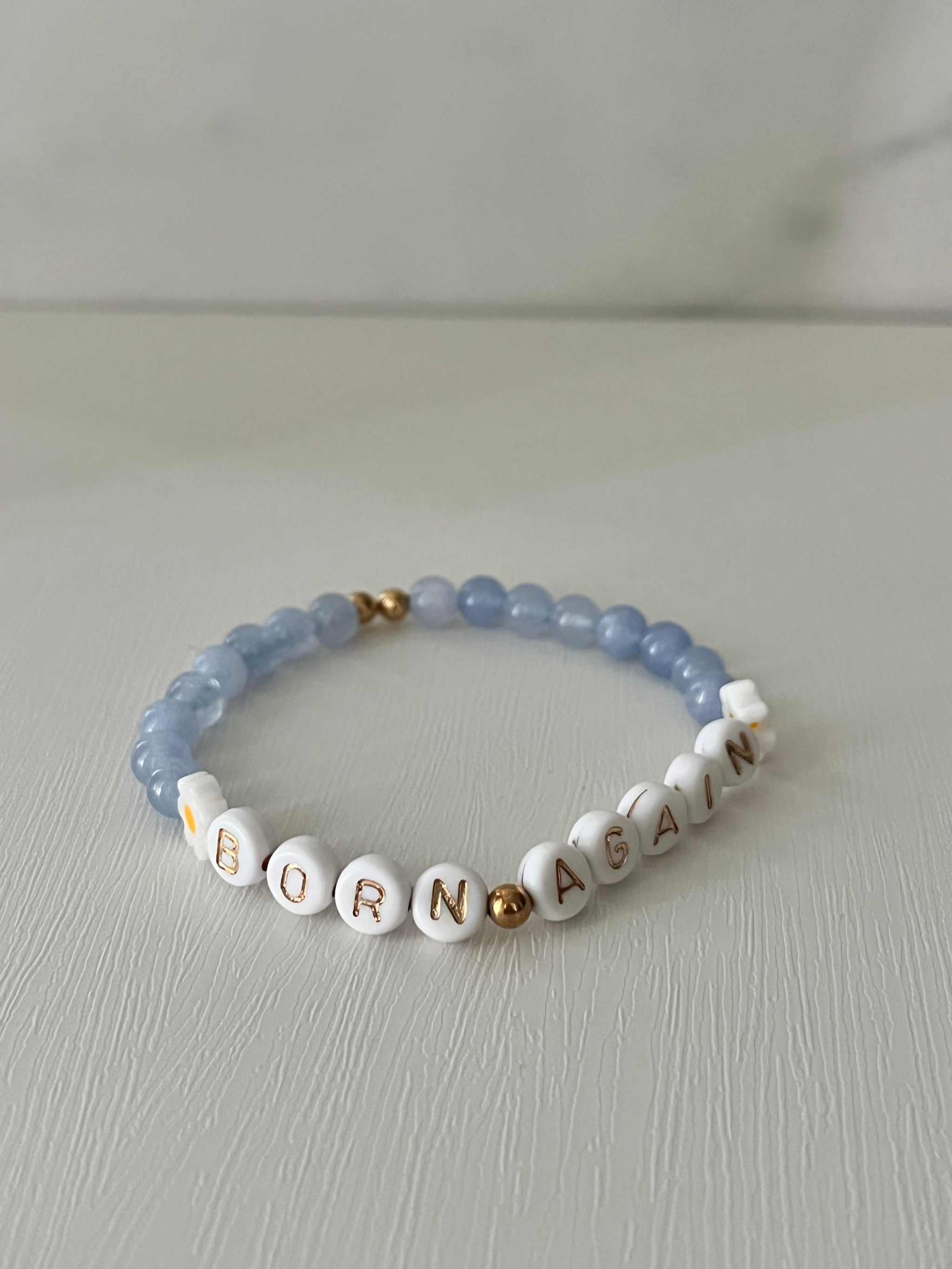 Sky blue beaded bracelet with white alphabet beads spelling “BORN AGAIN” and gold spacer accents.