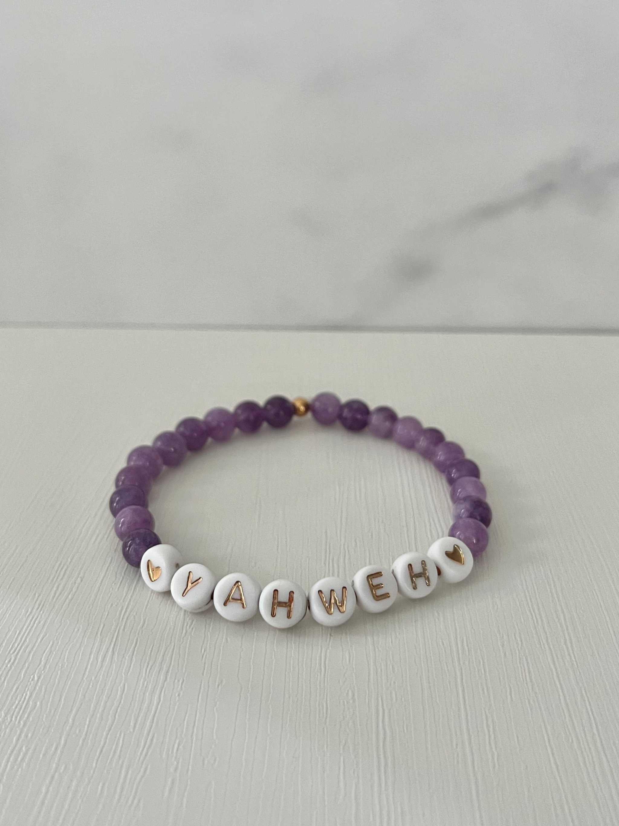 Purple beaded stretch bracelet with white letter charms spelling YAHWEH and small heart accents.