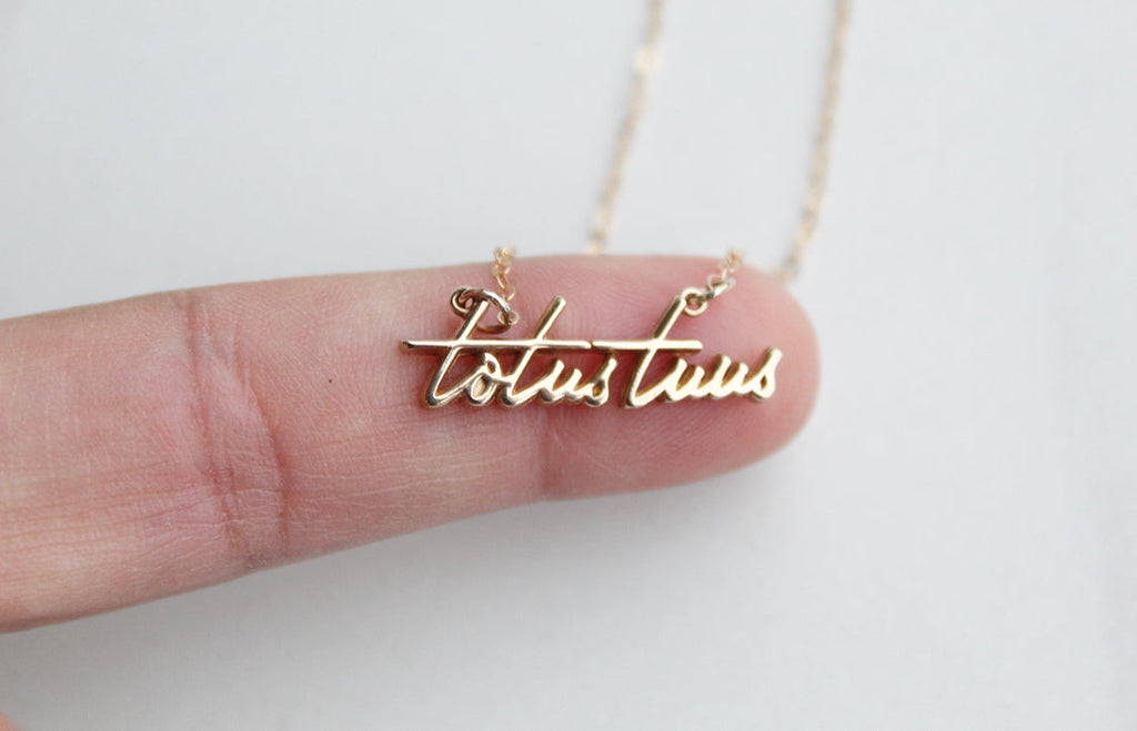 Gold-tone Totus Tuus script pendant necklace held in a hand