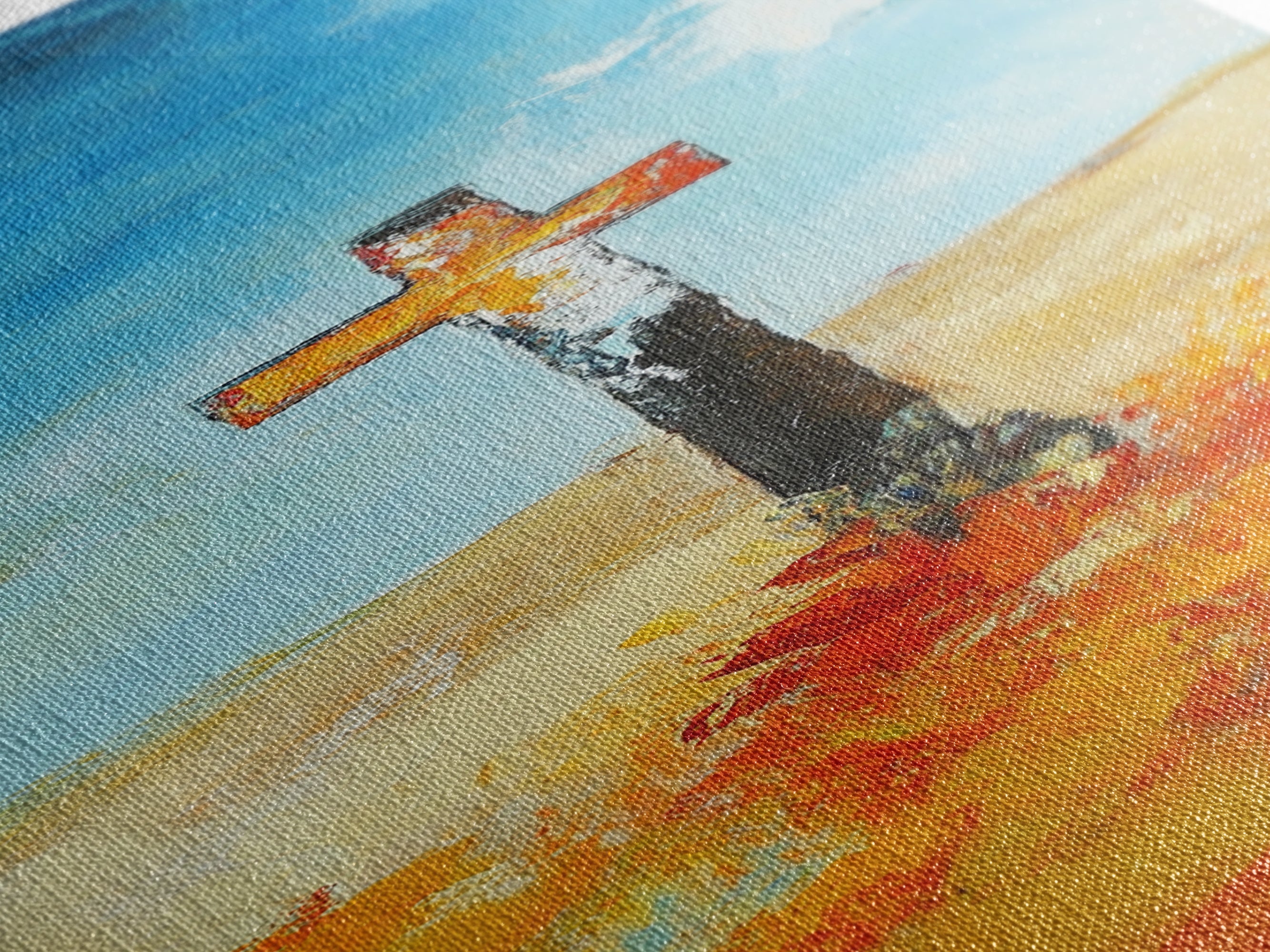 Textured cross painting close-up with blue sky, tan ground, and orange brushstrokes