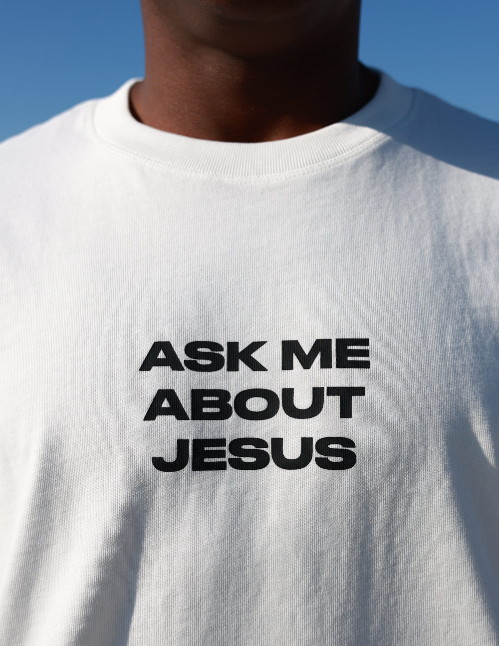 White heavyweight T-shirt close-up showing black text: Ask Me About Jesus.