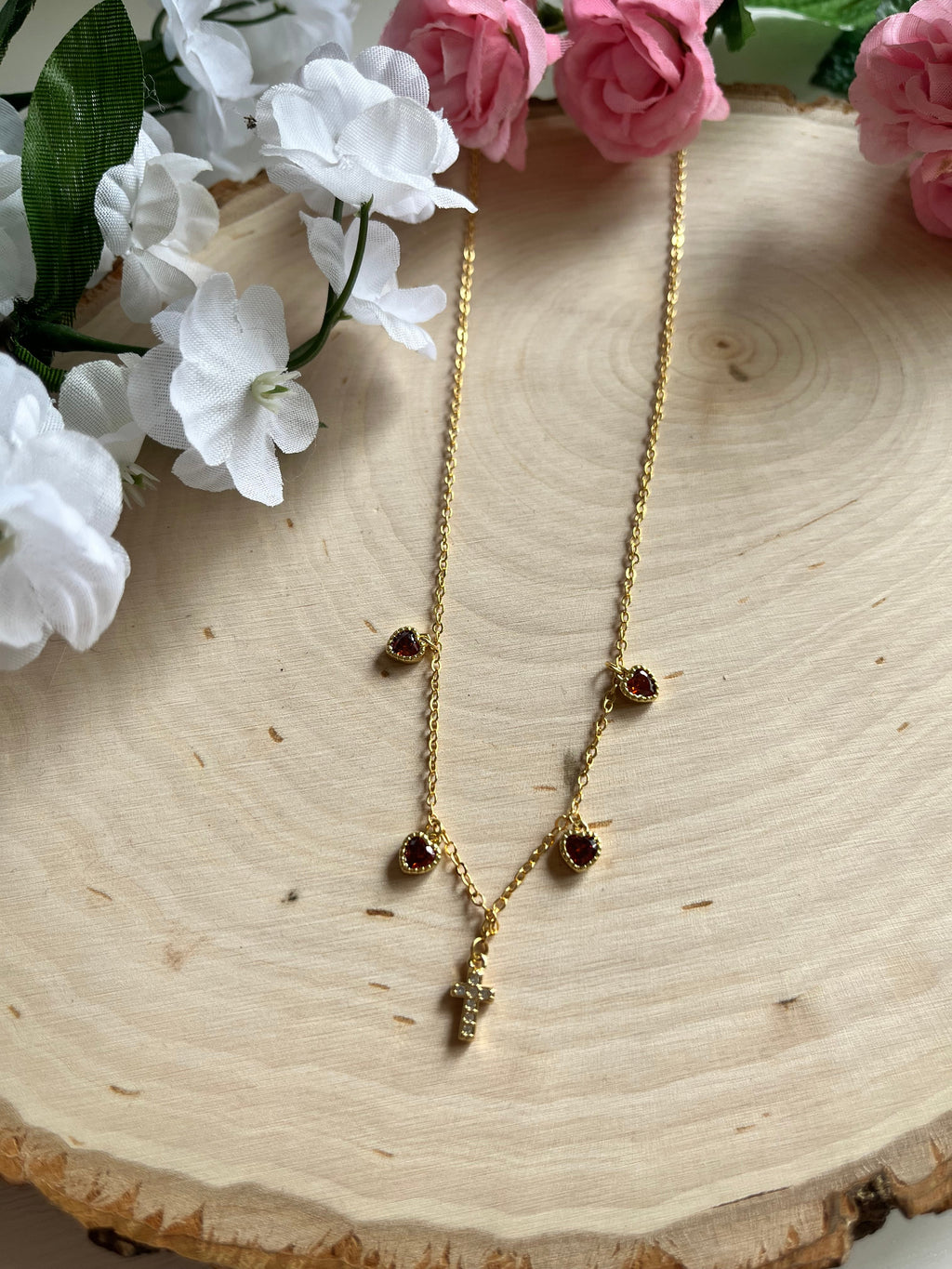 Gold charm necklace with red heart stones and gold cross pendant, angled view