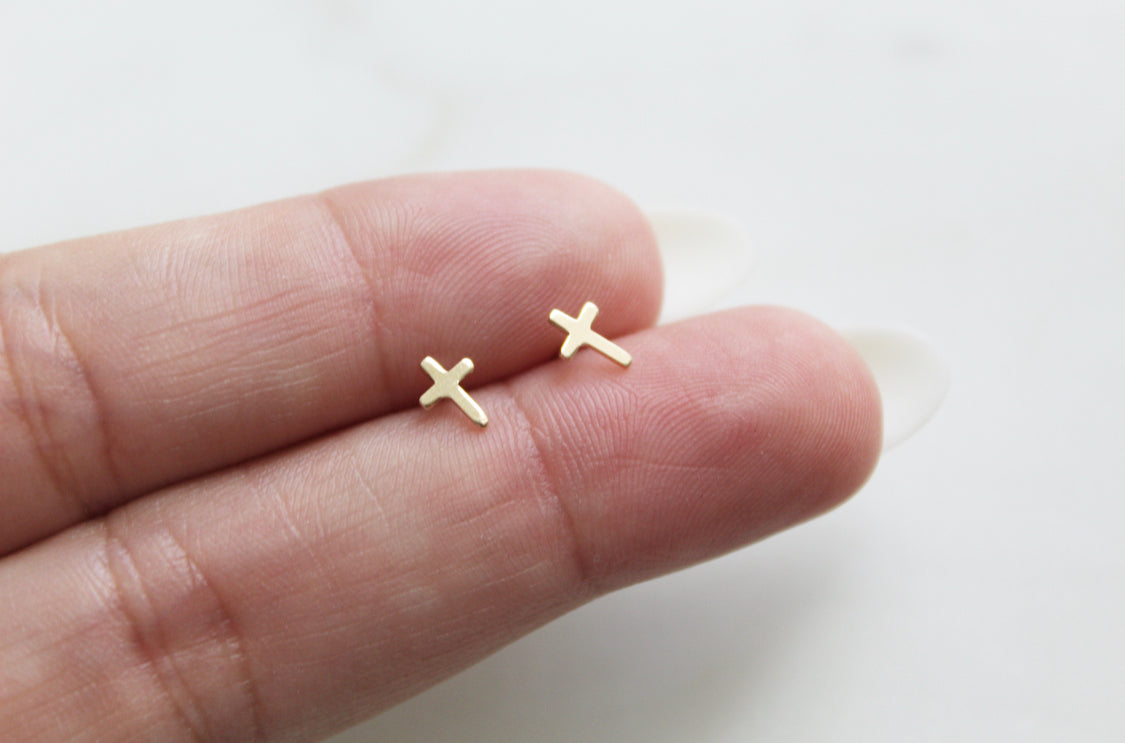 Pair of 14k gold cross stud earrings on fingertips, small cross shapes