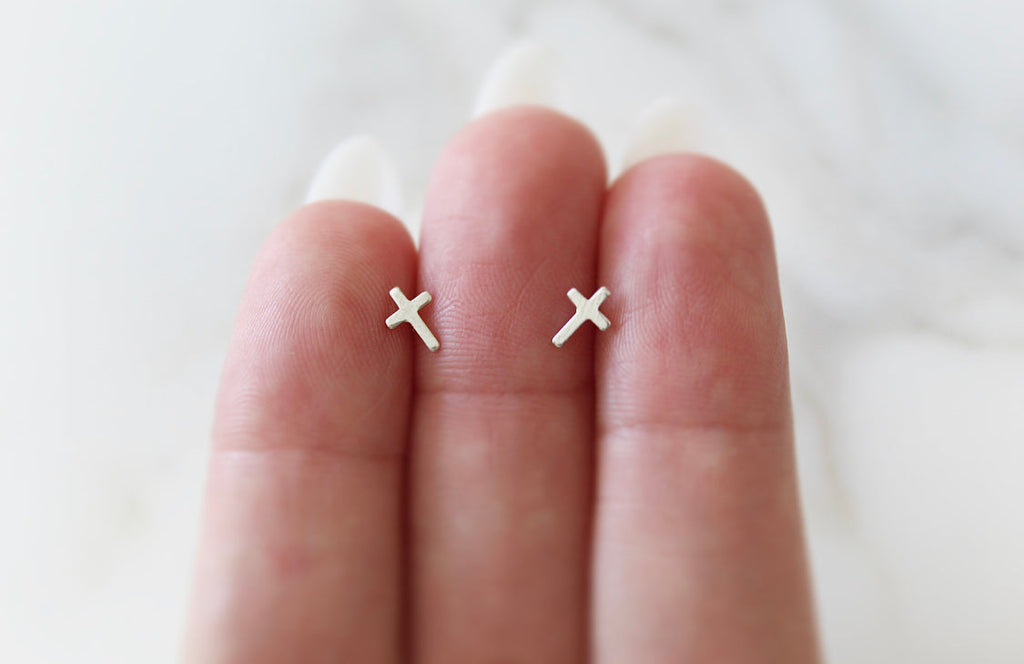 Sterling silver cross stud earrings on fingertips, two small cross charms visible.