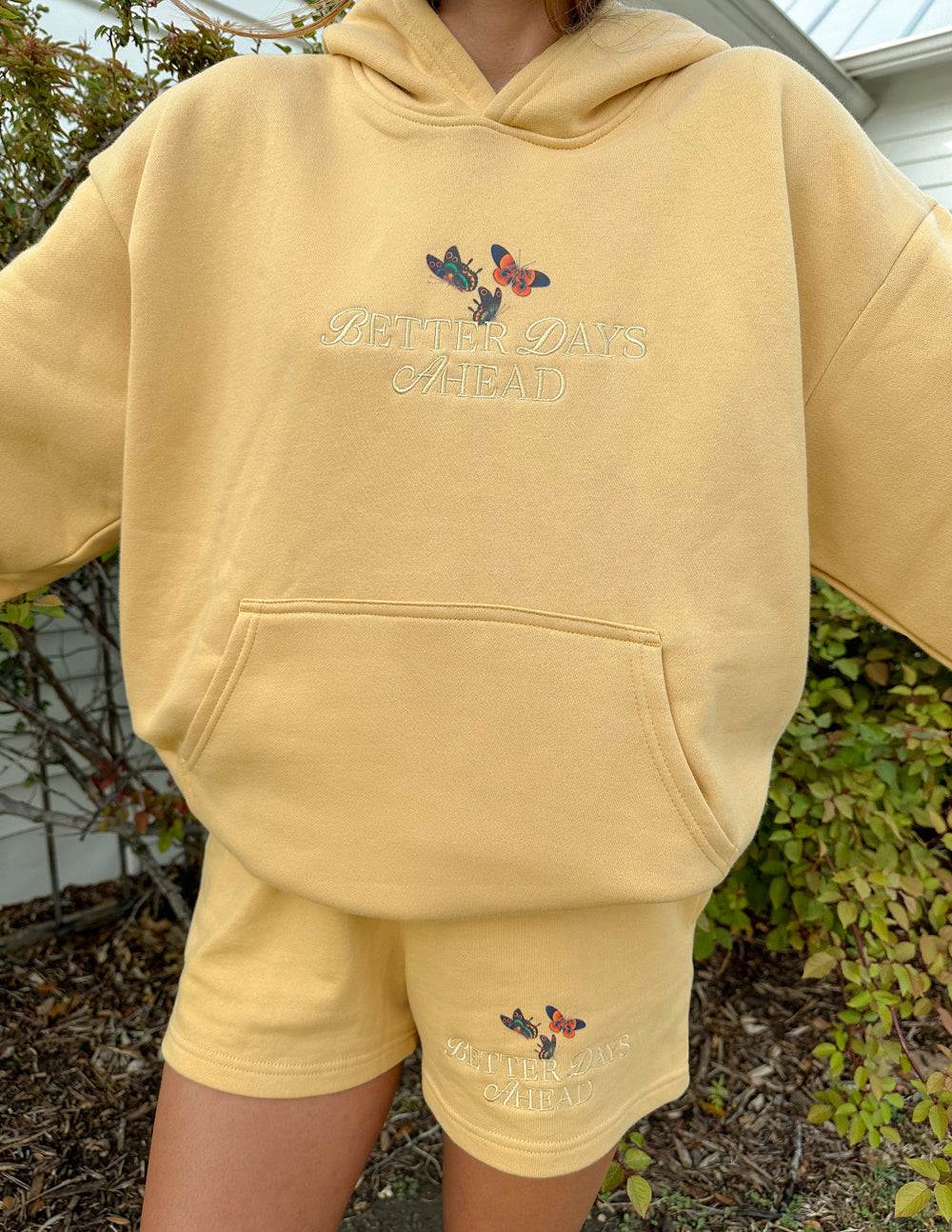 Unisex tan hoodie with butterfly logo on chest and kangaroo pocket, worn with shorts