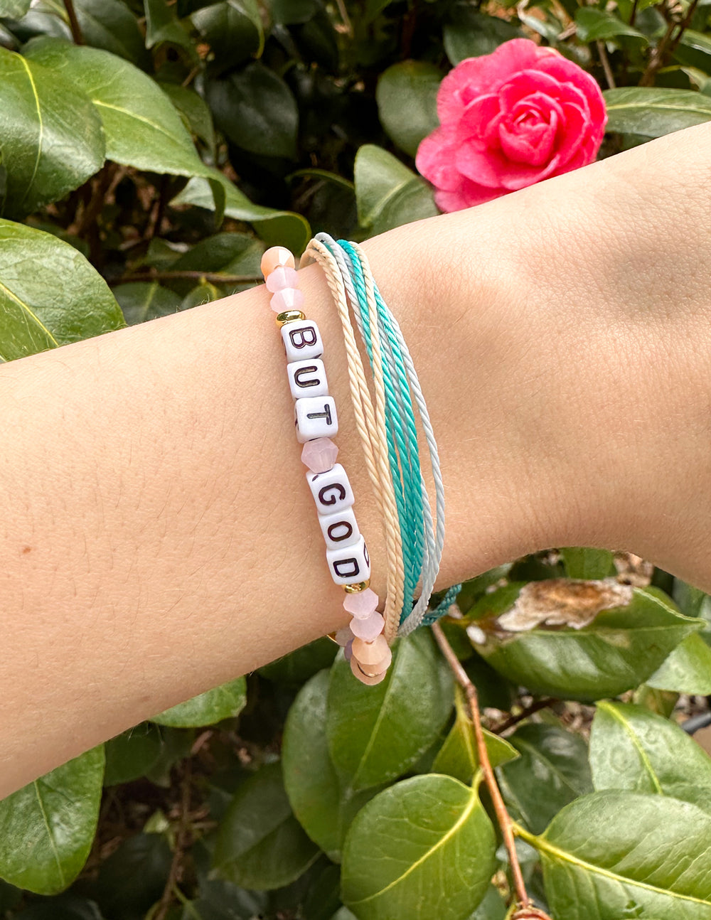 Beaded letter bracelet on wrist reading “BUT GOD”, paired with teal and white woven bands.