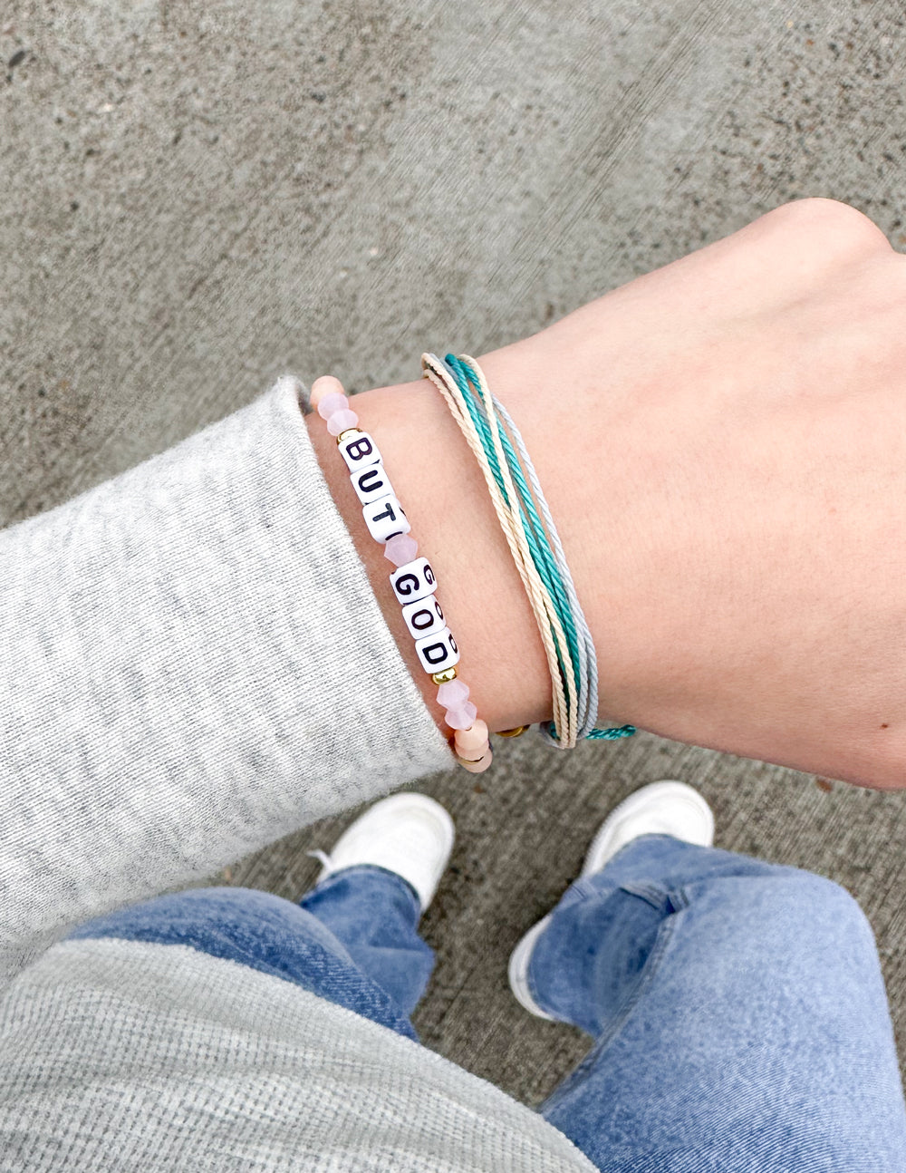 Stacked bracelets on wrist: beaded “BUT GOD” band with peach beads and teal cord wrap.