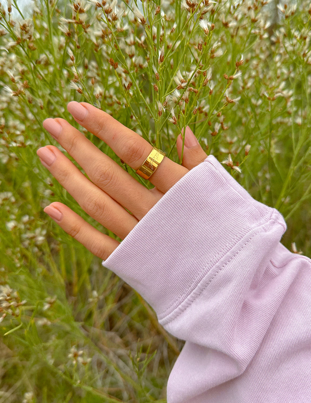 Gold engraved band ring on hand above grass; lavender sleeve visible