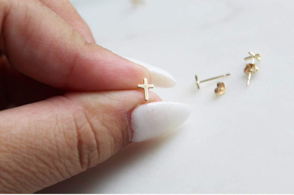 Gold-tone cross stud earrings with post backs, held beside an open palm.