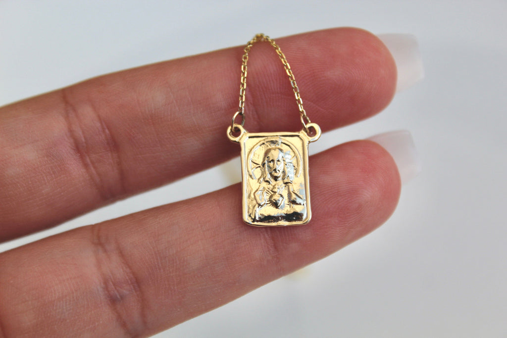 Gold scapular pendant held in hand, square front with figure relief on chain