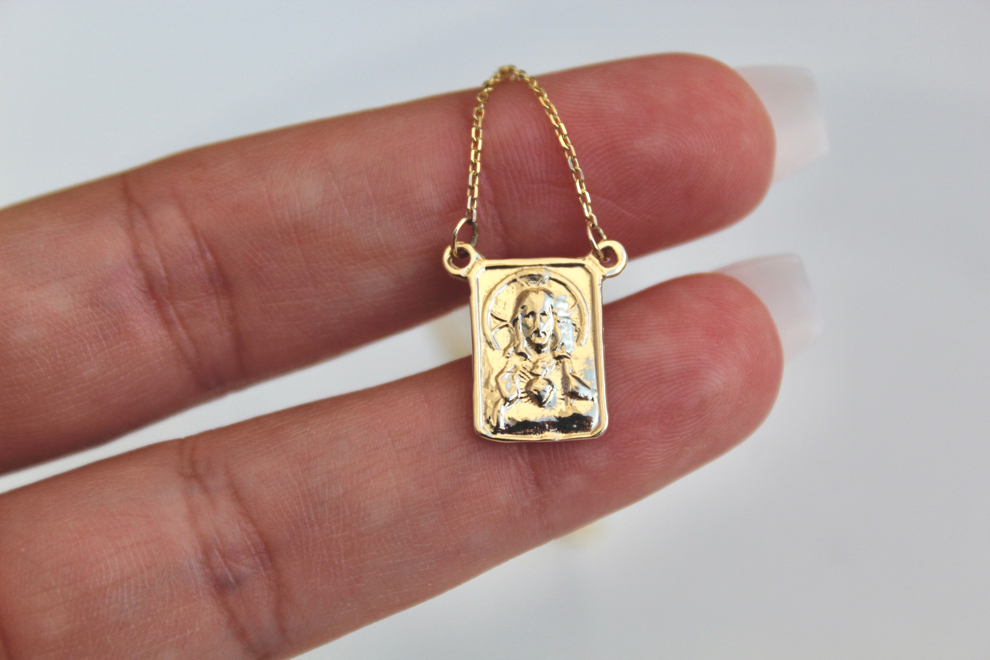 Gold scapular pendant held in hand, square front with figure relief on chain